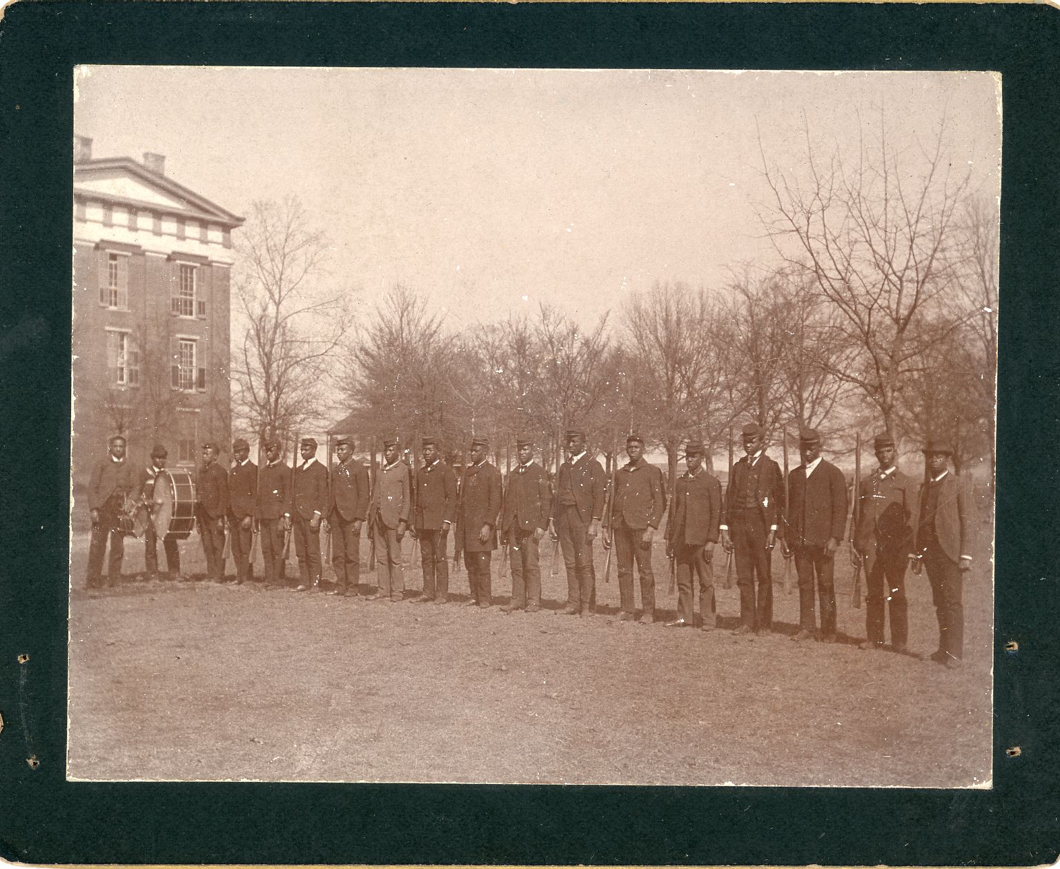 A sepia-tone photo of a straight line of African American men, wearing suits, standing in a field in front of a brick building.